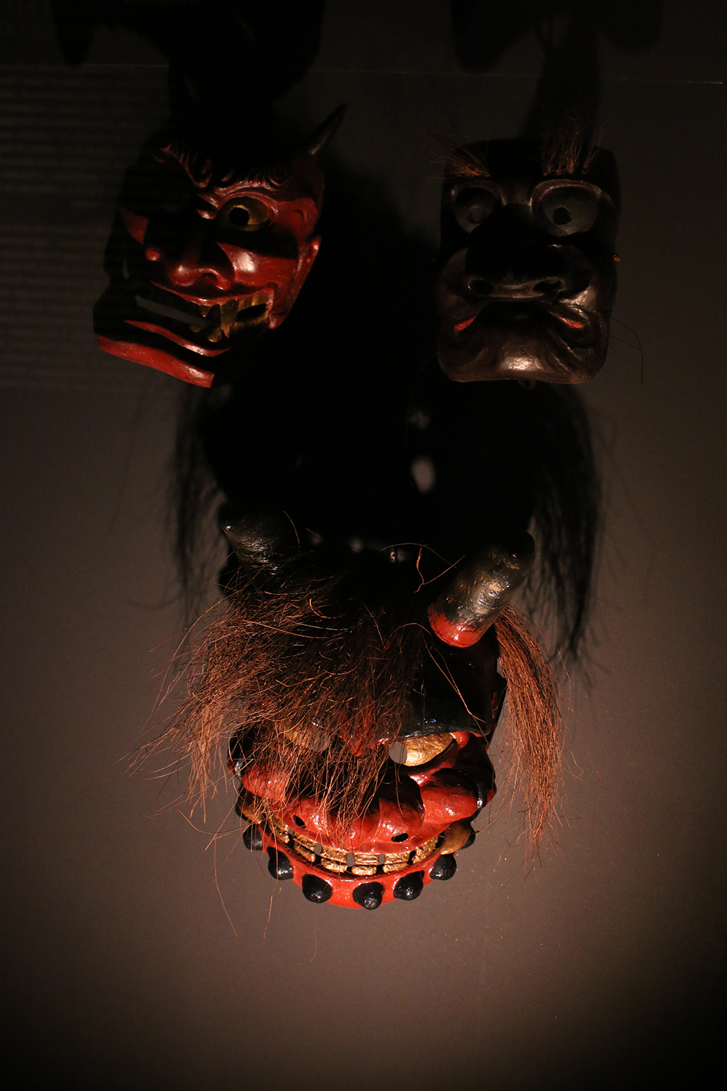"Demos", fotografía tomada no Museu do Oriente, Lisboa, o 1-6-23. Mostra unhas máscaras de oni (ou demos) do teatro xaponés, nunha exposición temporal sobre os rituais nipóns. Son tres máscaras vermellas de cabelo lacio e roxo e sorrisos arrepiantes, con luz dura dende abaixo sobre fondo escuro.