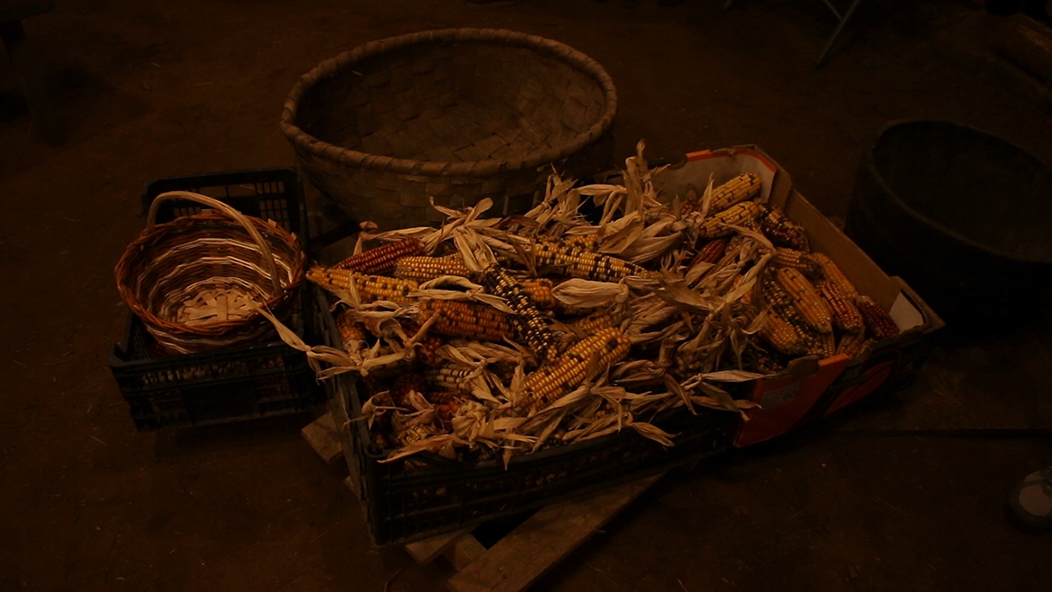 "Millo", fotografía tomada durante o rodaxe de O Trinxidoiro en Penelas, o 4-3-23. Mostra un cesto e varias caixas, a maior delas ten espigas de millo agardando a ser esfollado, cunha luz ténue e cálida.